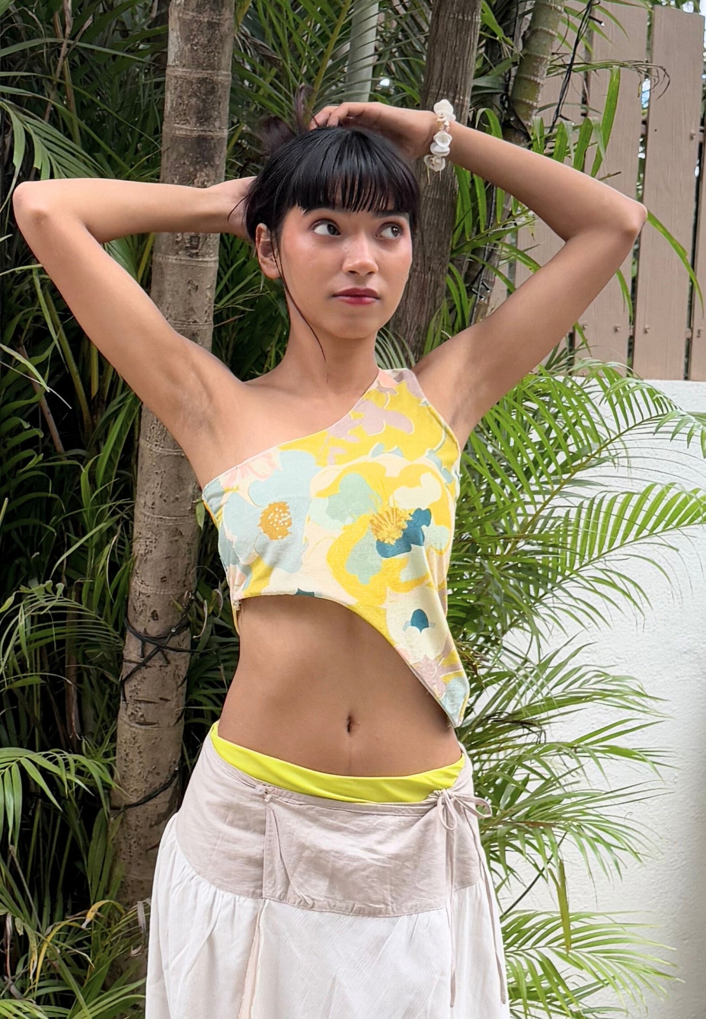 Woman wearing a colorful one-shoulder top and white pants in a tropical setting